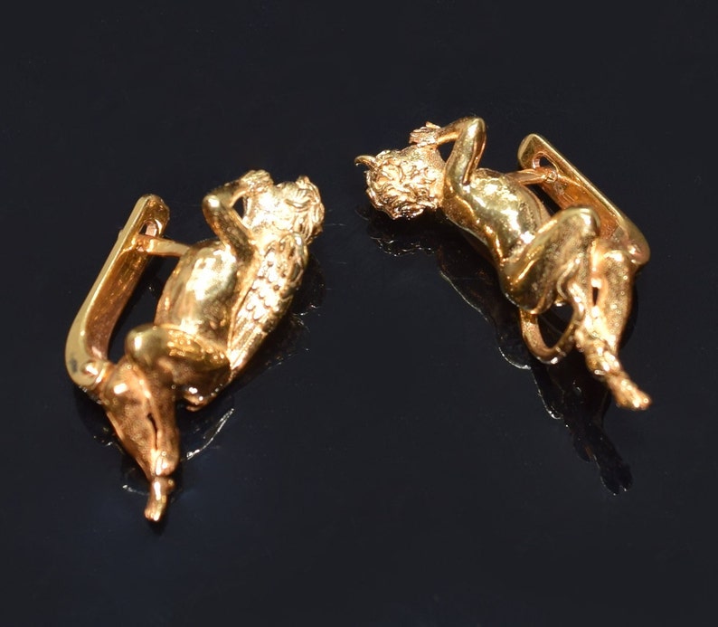 May include: A pair of gold-tone earrings featuring a cherub design. Each earring has a cherub figure with wings, lying down and holding its head in its hand.