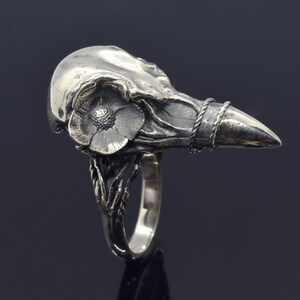 Puede incluir: Un anillo de plata con un dise&ntilde;o detallado de calavera de cuervo. Una flor est&aacute; esculpida en el lateral de la calavera, y un detalle de cuerda trenzada rodea el pico. La banda del anillo tiene un dise&ntilde;o org&aacute;nico texturizado.