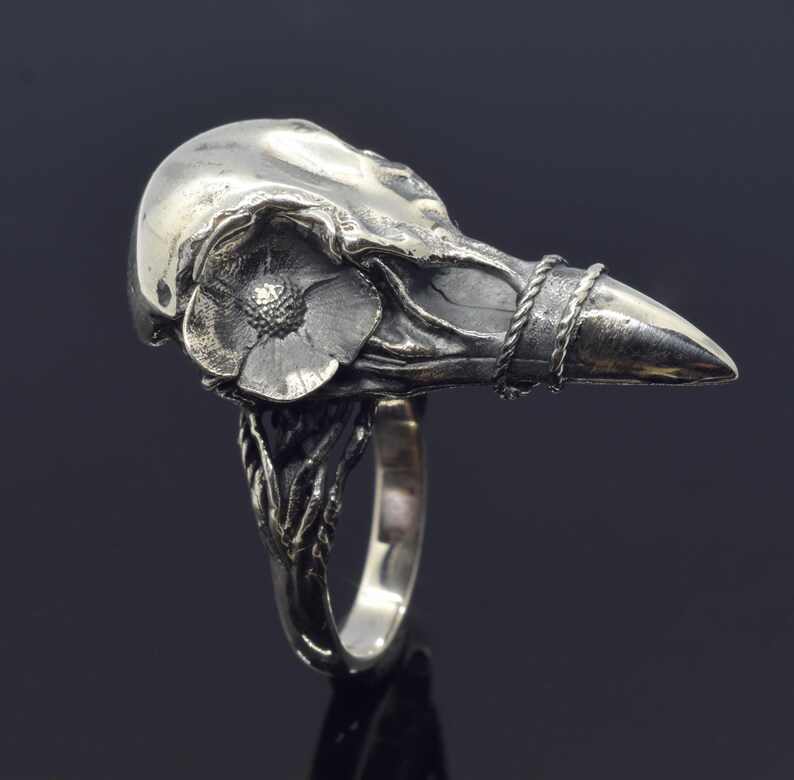 Puede incluir: Un anillo de plata con un dise&ntilde;o detallado de calavera de cuervo. Una flor est&aacute; esculpida en el lateral de la calavera, y un detalle de cuerda trenzada rodea el pico. La banda del anillo tiene un dise&ntilde;o org&aacute;nico texturizado.