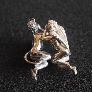 May include: A silver sculpture of an angel and a devil intertwined. The angel has wings and the devil has horns. The sculpture is made of metal and has a polished finish.