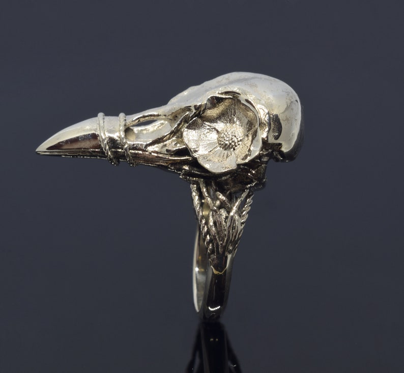 Puede incluir: Un anillo plateado con un dise&ntilde;o detallado de calavera de cuervo. El anillo incluye un adorno floral y detalles intrincados en la banda. La calavera de cuervo es el punto focal, con un pico puntiagudo y superficie texturizada.