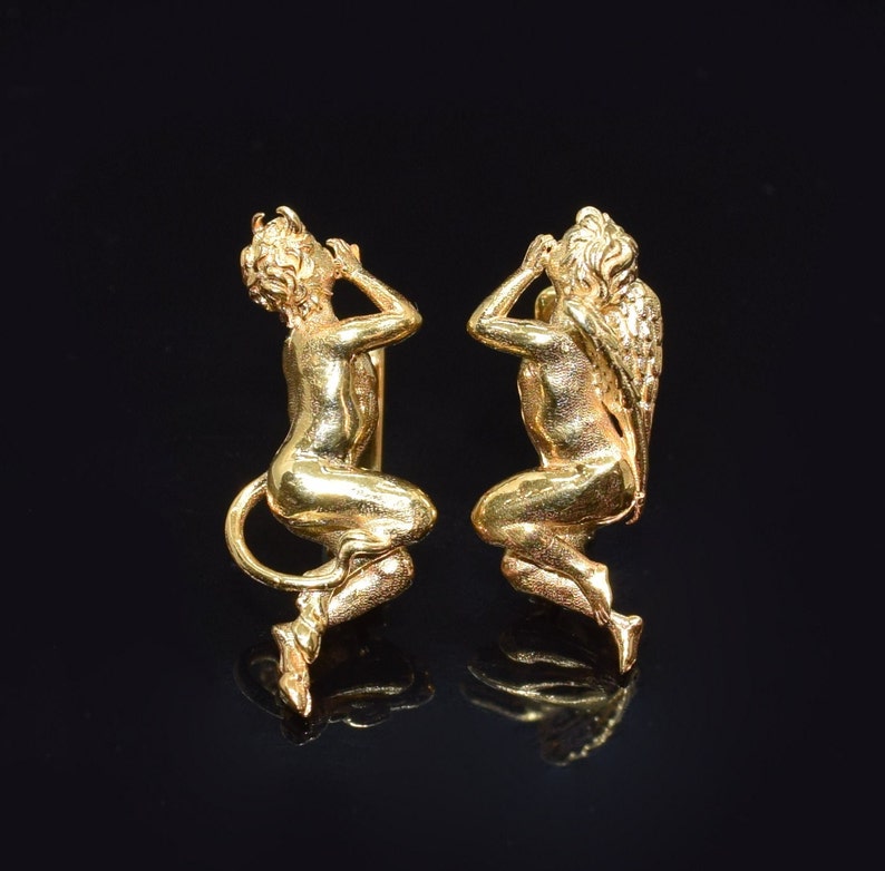 May include: A pair of gold-tone earrings featuring a cherub design. Each earring depicts a cherub with wings, sitting with one hand covering its face.