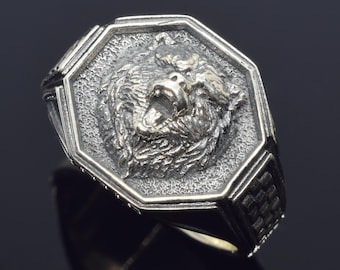 Könnte beinhalten: Silberfarbener Ring mit detailliertem Bärenkopf-Design. Das Gesicht des Bären befindet sich in einem achteckigen Rahmen. Der Ring hat eine strukturierte Oberfläche und ein kariertes Muster an den Seiten. Ein auffälliges Schmuckstück.