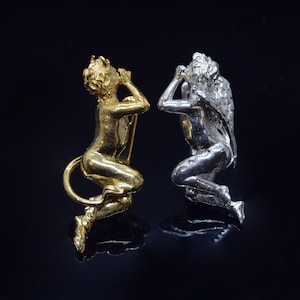 Peut inclure: Deux petites figurines d'anges détaillées, l'une dorée et l'autre argentée, avec des ailes et une main couvrant leur visage. Les figurines sont posées dans une position similaire, la tête penchée en arrière et les ailes déployées.