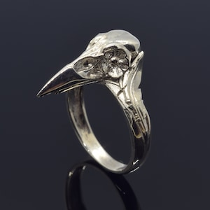 May include: A silver-colored ring featuring a detailed raven skull design. The ring's band is smooth, leading up to the intricately carved skull with a flower accent. The raven's beak is sharply pointed, and the ring has a polished finish.