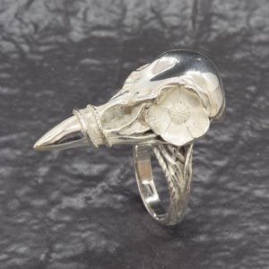 Puede incluir: Anillo plateado con dise&ntilde;o de calavera de p&aacute;jaro y una flor. La banda del anillo tiene una apariencia texturizada, similar a una vid. El pico de la calavera est&aacute; envuelto con un detalle de cuerda trenzada.