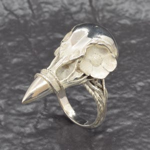 Puede incluir: Anillo de plata con dise&ntilde;o de calavera estilizada, punta y detalles florales. El anillo tiene una superficie texturizada y un acabado pulido. La calavera est&aacute; adornada con una flor. El dise&ntilde;o es intrincado y detallado.