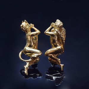 May include: A pair of gold-toned earrings featuring a detailed sculpture of a cherub with wings on one side and a cherub with a tail on the other.