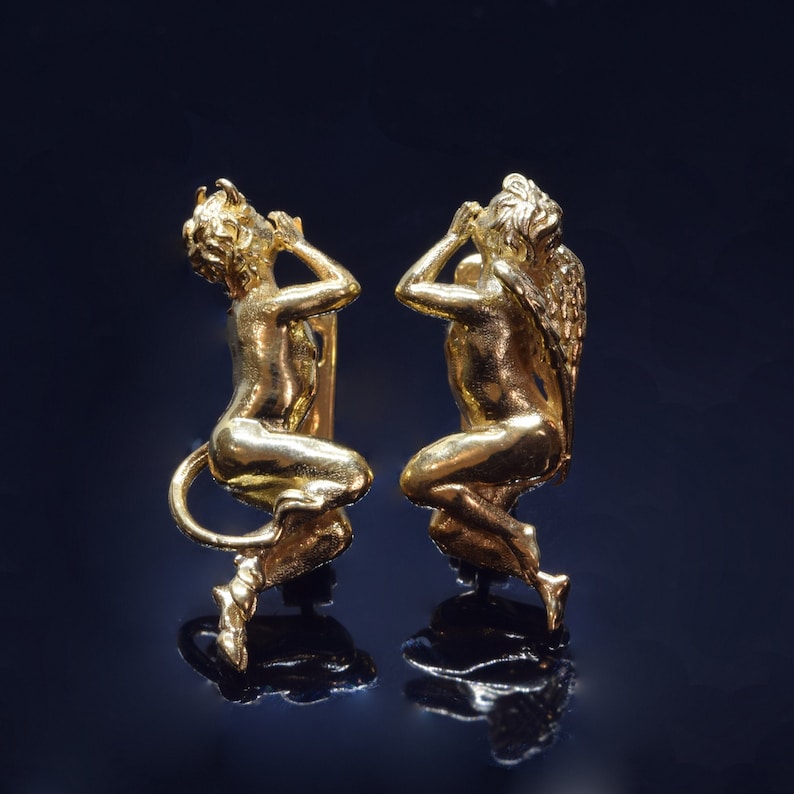 May include: A pair of gold-toned earrings featuring a detailed sculpture of a cherub with wings on one side and a cherub with a tail on the other.