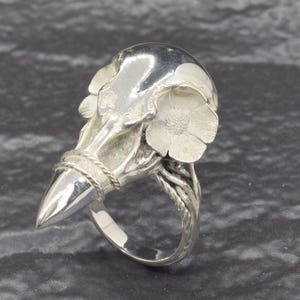 Puede incluir: Un anillo de plata con un dise&ntilde;o de calavera de p&aacute;jaro estilizado. El anillo tiene un pico puntiagudo, detalles florales y un detalle de cuerda trenzada alrededor de la base del pico. La banda del anillo es lisa y pulida.