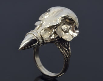 May include: A silver ring with a skull and flower design. The ring has a pointed, beak-like extension and detailed band. The skull's eye socket is visible, and the flower is in full bloom. The ring is a unique piece of jewellery.