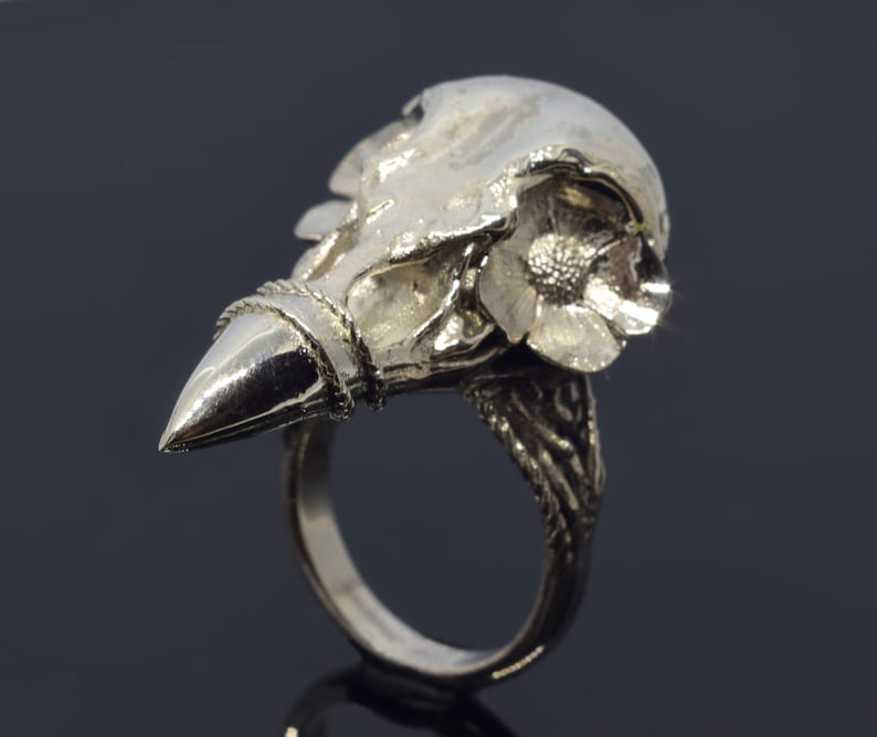 Puede incluir: Anillo plateado con dise&ntilde;o de calavera y flor. El anillo tiene una extensi&oacute;n puntiaguda en forma de pico y una banda detallada. La cuenca del ojo de la calavera es visible y la flor est&aacute; en plena floraci&oacute;n.