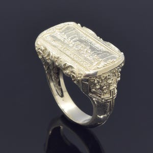 Può includere: Anello color argento con una superficie rettangolare con un design a rilievo dettagliato. L'anello presenta volute ornate sui lati e un disegno a croce sullo stelo. L'anello è posto su uno sfondo scuro.