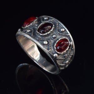 Pode incluir: Um anel de prata com tr&ecirc;s pedras preciosas vermelhas engastadas em um design detalhado. O anel tem uma banda larga e uma superf&iacute;cie texturizada.