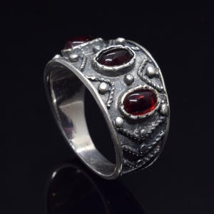 Pode incluir: Um anel de prata com um design intrincado e tr&ecirc;s pedras preciosas vermelhas. O anel tem uma banda larga e uma superf&iacute;cie texturizada.