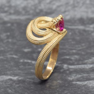 May include: A gold ring shaped like a coiled snake with a textured, beaded surface. The snake head is positioned at the top, featuring a small, oval-shaped, vibrant pink gemstone set within its open mouth, creating a detailed and ornate jewelry piece.