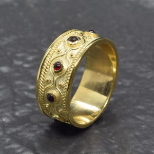 May include: A gold ring with a detailed pattern and red gemstones. The ring has an ornate design with swirls and dots. The band features several small, round, red gemstones. The ring is displayed on a dark surface.