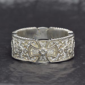 Könnte beinhalten: Ein silberner Ring mit einem komplexen Reliefmuster aus sich wiederholenden Kreuzmotiven und kunstvollen floralen Ranken. Das Band hat eine texturierte, oxidierte Oberfläche, die die detaillierten Gravuren auf der gesamten Außenseite hervorhebt.