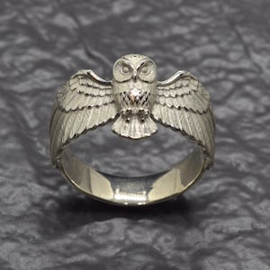 May include: A silver-toned metal ring featuring a detailed owl with outstretched wings. The owl is centred on the band, with its wings extending along the sides. The metal has a textured finish on the owl's feathers and a smooth, polished band.