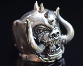 May include: A silver ring with a detailed design of a snarling, horned creature with a chain and a nose ring. The ring is made of metal and has a textured surface.