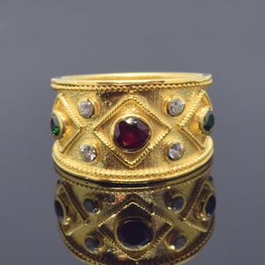 Op de afbeelding: Een gouden ring met een brede band met een geometrisch ontwerp met rode, groene en witte edelstenen.