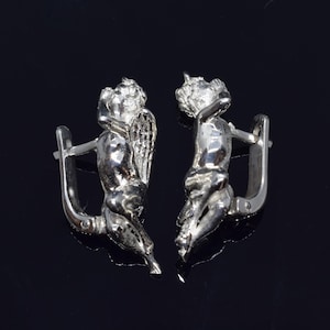 May include: A pair of silver earrings featuring a detailed design of a cherub with wings. The earrings are made of a shiny metal and have a hook closure.