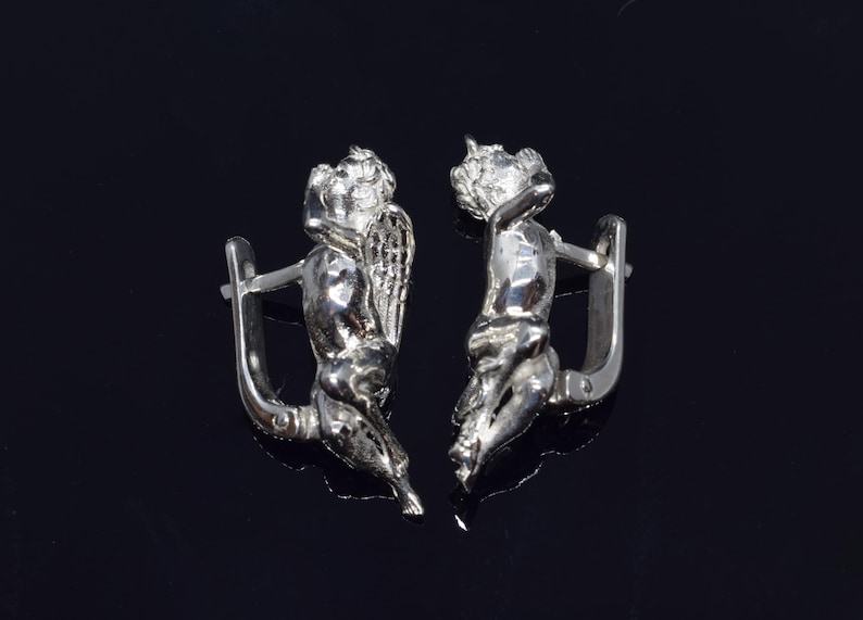 May include: A pair of silver earrings featuring a detailed design of a cherub with wings. The earrings are made of a shiny metal and have a hook closure.