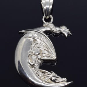 May include: A silver pendant featuring a detailed dragon curled around a crescent moon. The dragon has wings and a long tail, and its head is turned to the side. The pendant is attached to a silver bail.