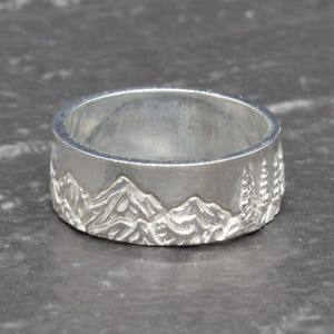 May include: A silver ring with a mountain range design. The ring features a detailed relief of mountains and trees around the band. The ring is made of a shiny, metallic material.