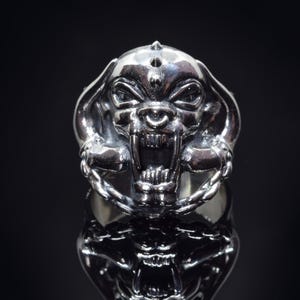May include: A silver ring with a detailed design of a snarling wolf's head with sharp teeth and a chain around its neck.