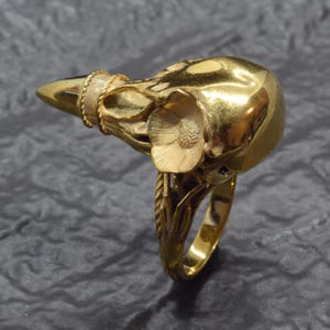Puede incluir: Un anillo dorado con forma de cr&aacute;neo de ave, con detalles intrincados. El anillo tiene una banda texturizada alrededor del pico y un acento floral cerca de la cuenca del ojo. La banda del anillo tiene un dise&ntilde;o similar a una cuerda.