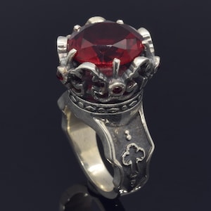 May include: A silver ring with a large, red gemstone set in a crown-shaped design. The ring band features intricate details and a cross.