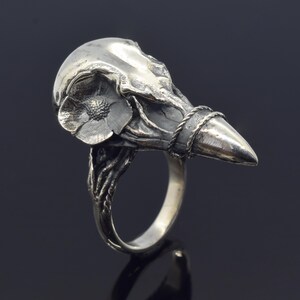 Puede incluir: Un anillo plateado con un dise&ntilde;o de calavera de p&aacute;jaro estilizado. El anillo tiene un detalle floral en la calavera y una banda trenzada alrededor del pico. El anillo est&aacute; sobre un fondo oscuro.