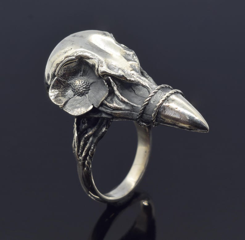 Puede incluir: Un anillo plateado con un dise&ntilde;o de calavera de p&aacute;jaro estilizado. El anillo tiene un detalle floral en la calavera y una banda trenzada alrededor del pico. El anillo est&aacute; sobre un fondo oscuro.