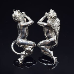 May include: A pair of silver cherub figurines, one with wings and one with a tail, both with their hands covering their eyes.
