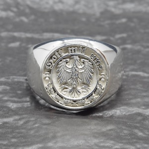 May include: A silver ring with a detailed emblem featuring an eagle and the words "Gott mit uns" in a circular design. The ring has a polished, reflective surface and a wide band.