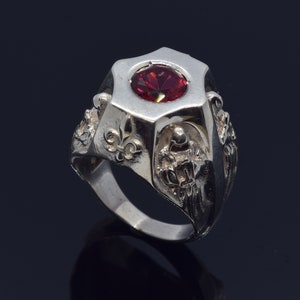 Könnte beinhalten: Ein silberner Ring mit einem großen, facettierten, ovalen, roten Edelstein. Der Ring hat ein geometrisches Design um den Stein und verzierte Details am Band, einschließlich Fleur-de-Lis und anderer skulpturaler Elemente. Der Ring ist vor dunklem Hintergrund platziert.