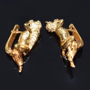 May include: A pair of gold-toned earrings featuring a detailed cherub design. The cherubs are depicted in a reclining pose with wings and intricate details.