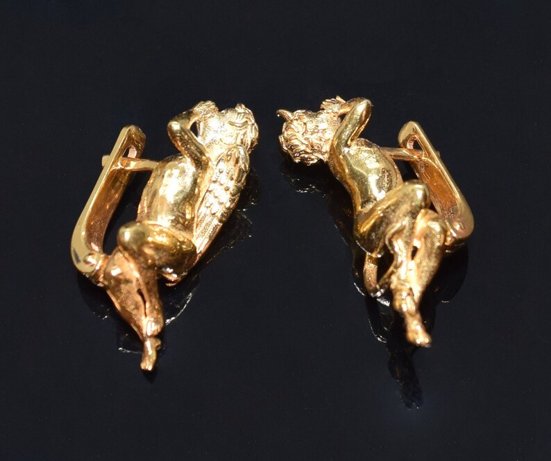 May include: A pair of gold-toned earrings featuring a detailed cherub design. The cherubs are depicted in a reclining pose with wings and intricate details.
