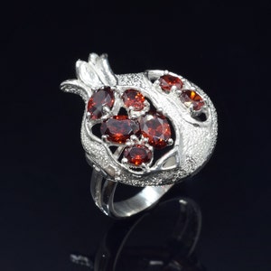 Può includere: Un anello d'argento a forma di melograno con pietre preziose rosse.