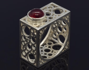 May include: A silver ring with a geometric design featuring a square shape with cut-out circles. A round, red gemstone is set on top. The ring has a modern, artistic style.