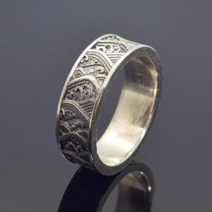 Może przedstawiać: Srebrny pierścionek z detalicznym wzorem fal. Pierścionek jest wypolerowany i ma gładkie wykończenie.