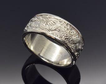 Può includere: Un anello d'argento con un design dettagliato di drago che avvolge la fascia. L'anello ha un interno lucido e un esterno strutturato, che mostra le squame e le caratteristiche del drago. L'anello è esposto su una superficie riflettente.