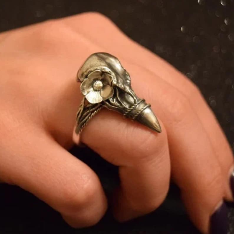 Puede incluir: Un anillo plateado con un dise&ntilde;o de calavera de p&aacute;jaro estilizado. Una peque&ntilde;a flor est&aacute; colocada en la parte superior de la calavera. La banda del anillo tiene una textura tejida. El anillo se lleva en un dedo.