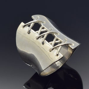 May include: A silver ring with a corset-like design. The ring is made of smooth, polished metal and features a criss-cross pattern of raised lines.