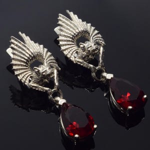 May include: Silver earrings with a bat design and a dangling red teardrop-shaped gemstone.