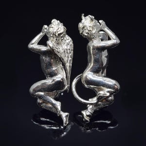 May include: A pair of silver cherub figurines with wings. The cherubs are facing each other and have their hands raised to their faces.