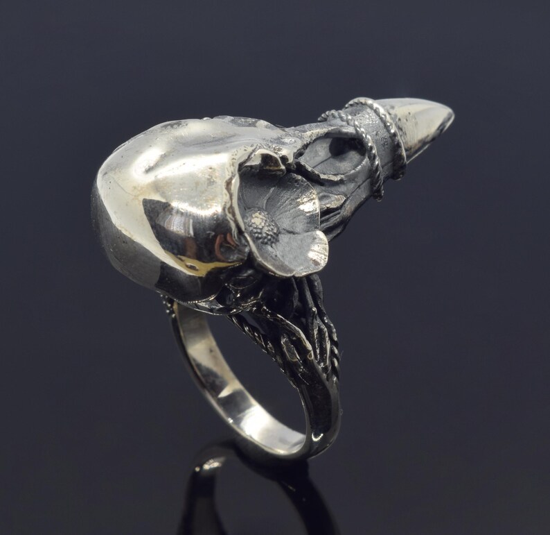 Puede incluir: Anillo plateado con dise&ntilde;o de calavera, con una flor y un elemento puntiagudo. El anillo presenta detalles intrincados y un acabado pulido, sobre un fondo oscuro. Una joya &uacute;nica con dise&ntilde;o de calavera.