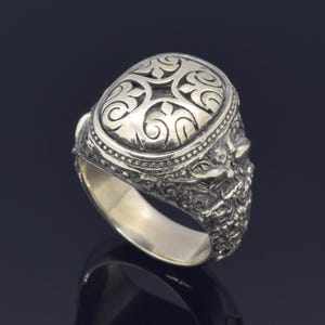May include: A silver ring with an intricate design. The top of the ring features a circular, raised design with a floral pattern. The band of the ring is also decorated with a detailed floral pattern.