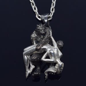 May include: A silver pendant necklace featuring a detailed sculpture of a wolf holding a woman in its arms. The pendant is made of two different metals, with the wolf being a darker silver and the woman being a lighter silver.